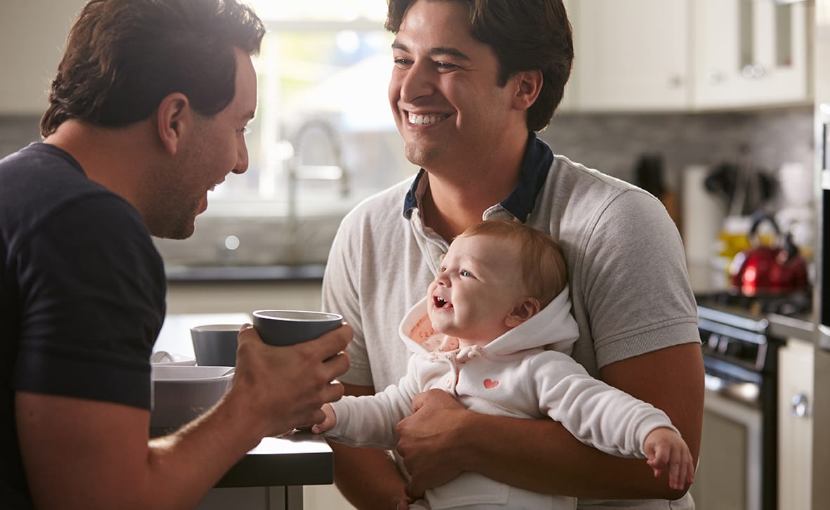 Two men in a kitchen holding a baby